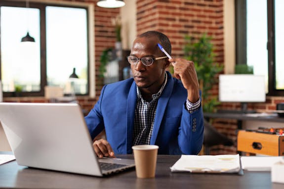 A business man in a blue suit holding a pen to his head and sitting in front of a laptop that is beside a coffee cup.